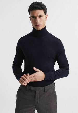Pulover de tip guler înalt, de culoare bleumarin, realizat dintr-un material tricotat neted, cu un design ajustat și mâneci lungi. Este purtat cu pantaloni eleganți maro, iar mâinile sunt poziționate în față.