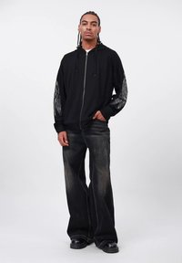 Uomo con capelli intrecciati che indossa una felpa nera con zip e motivi sulle maniche, jeans neri a gamba larga sbiaditi e scarpe nere, in piedi contro uno sfondo bianco.