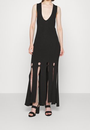 Schwarzes ärmelloses Kleid mit tiefem V-Ausschnitt, seitlichen Schlitzen und ausgeschnittenen Akzenten am Saum. Kombiniert mit schwarzen High-Heels.