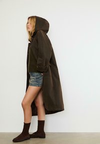 Donna che indossa un lungo cappotto marrone con cappuccio, un maglione verde, pantaloni shorts in denim, calzini marroni e mocassini marroni, in piedi di lato contro un muro semplice.