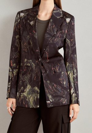 Mujer que lleva un blazer oscuro con patrones abstractos en verde y naranja, combinado con una camiseta oscura y pantalones negros sueltos con bolsillos.