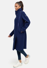 Manteau long navy avec col haut, poches latérales, fermeture éclair avant intégrale et texture douce. Porté sur un jean bleu et des baskets blanches.