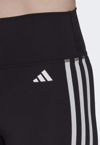 Czarne sportowe legginsy z wysokim stanem, z białym logo Adidas oraz trzema białymi paskami na boku. Gładka faktura i elastyczny materiał.