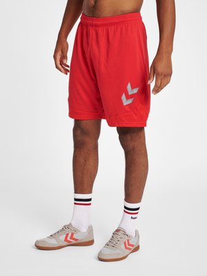 Mand iført røde atletikshorts med gråt Chevron-logo, hvide crew-sokker med sorte og røde striber og beige sneakers med orange chevroner.