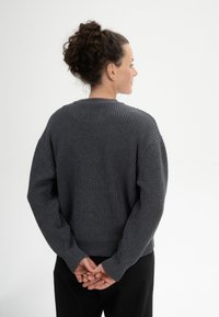 Gestrickter dunkelgrauer Pullover mit strukturiertem Ribbenmuster, weiten Ärmeln und rundem Halsausschnitt, rückwärts getragen mit hinter dem Rücken verschränkten Händen.