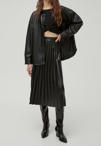 Giacca camicia in pelle nera, top corto, gonna midi plissettata e stivali con tacco fino al ginocchio, tutti con una finitura lucida, creando un look monocromatico coerente.
