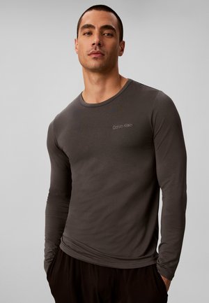 Mann trägt ein eng anliegendes dunkelgraues Calvin Klein Langarmshirt und steht mit den Händen in den Taschen vor einem einfarbigen hellen Hintergrund.