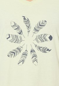 Světle zbarvené látkové tričko s kulatým designem stylizovaných černých peří, z nichž každé má složité vzory a detaily.