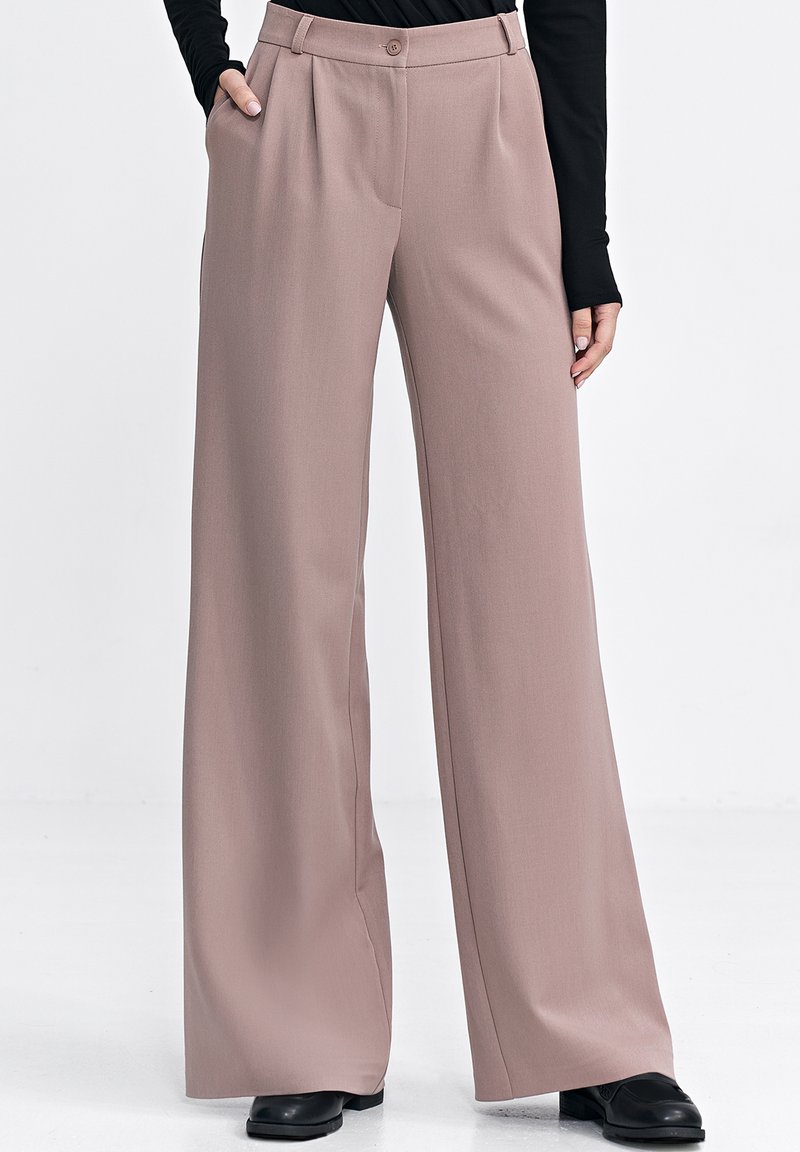 Jasnoróżowe spodnie o szerokich nogawkach o gładkiej fakturze, z zapięciem na guzik i pliskami. W połączeniu z czarnym obuwiem.