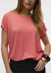 Koralfarvet kortærmet top med bådudskæring, lavet af blødt, struktureret stof, parret med mørke jeans og flere armbånd på håndleddet.