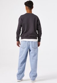 Sudadera gris oscura con puños y dobladillo acanalados, combinada con jeans sueltos de color azul claro. El modelo está de espaldas frente a un fondo blanco.