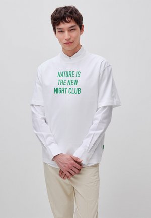 Młody mężczyzna w białej warstwowej koszuli z zielonym napisem „NATURE IS THE NEW NIGHT CLUB” i beżowych spodniach, stojący na jednolitym tle.