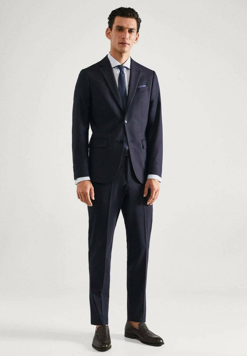 Costume marine à la coupe ajustée, avec une fermeture à un bouton et des revers crantés. Accompagné d'une chemise bleu clair et d'une cravate à motifs.