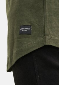 Olive green t-shirt with a curved hem, featuring a black label with white text "JACK & JONES EST. 1990" sewn at the lower side.