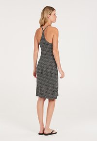 Vestido de patrón en blanco y negro con cuello halter, diseño ajustado, dobladillo hasta la rodilla y textura de tela suave. Lleva sandalias planas.