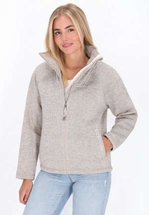 Jonge vrouw in een lichtgrijze fleece jas met rits en hoge kraag, draagt lichte blauwe spijkerbroek en glimlacht met één hand in haar zak.