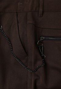 Pantalon en tissu marron avec une poche à fermeture éclair, un détail chaîne et des accents cousus. Le matériau est durable avec une texture lisse.