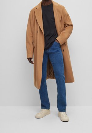 Osoba ubrana w długi, wielbłądzi płaszcz, czarną koszulę, niebieskie dżinsy oraz beżowe trampki, stojąca na tle jednolitego jasnoszarego tła.