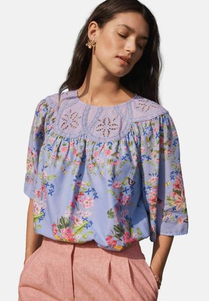 Kobieta o długich ciemnych włosach nosi lawendową bluzkę w kwiaty z koronkową wstawką i różowe spodnie z wysokim stanem, do których dobrała kolczyki w kształcie kwiatów.