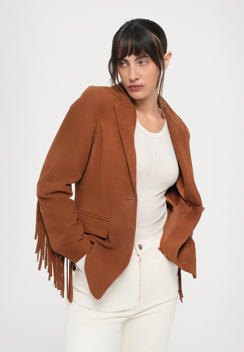 Blazer en suède marron avec des franges sur les manches, doté d'une fermeture à un seul bouton et de deux poches avant, porté sur un haut et un pantalon clairs.