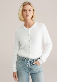 Weißer Cardigan aus weichem, strukturiertem Material. Mit sechs runden Knöpfen, einem runden Halsausschnitt und schmalen Ärmeln. Kombiniert mit hellblauen Jeans.
