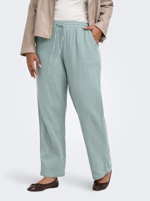 Pantaloni larghi verde chiaro con coulisse in tessuto strutturato, abbinati a scarpe basse marroni e una giacca beige sopra una maglia bianca.