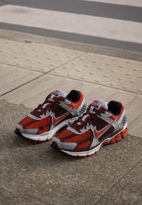 Röda och grå atletiska skor med mesh-sidor, vadderade sulor och framträdande Nike-logotyp. Har svarta och orangea accenter.