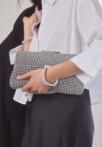 Vrouw draagt een wit shirt en een zwarte rok, met een zilveren clutch met strass steentjes en een zilveren armband en bangle.