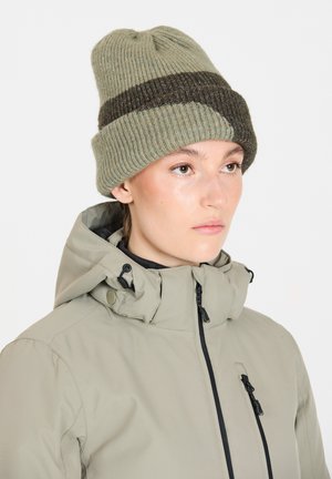 Stickad mössa i ljus olivgrön med en mörkbrun rand. Ribbad textur. Modellen bär en ljus jacka med hög krage och dragkedjor.