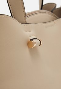 Beigefarbene Ledertasche mit glatter Textur, einem runden goldfarbenen Metallakzent und genähten Details entlang der Kanten und Nähte.