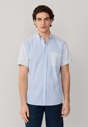 Junger Mann trägt ein kurzärmeliges Hemd mit hellblauen und weißen vertikalen Streifen, einer Brusttasche vorne und dunkle Hosen, steht vor einem einfarbigen Hintergrund.