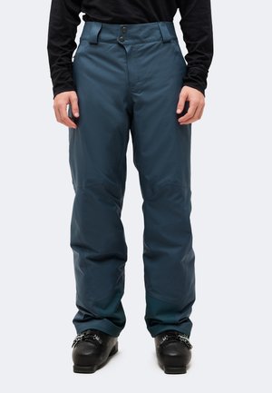 Le modèle porte un pantalon de ski bleu canard foncé avec une ceinture à deux boutons et des chaussures de ski noires sur un fond uni.