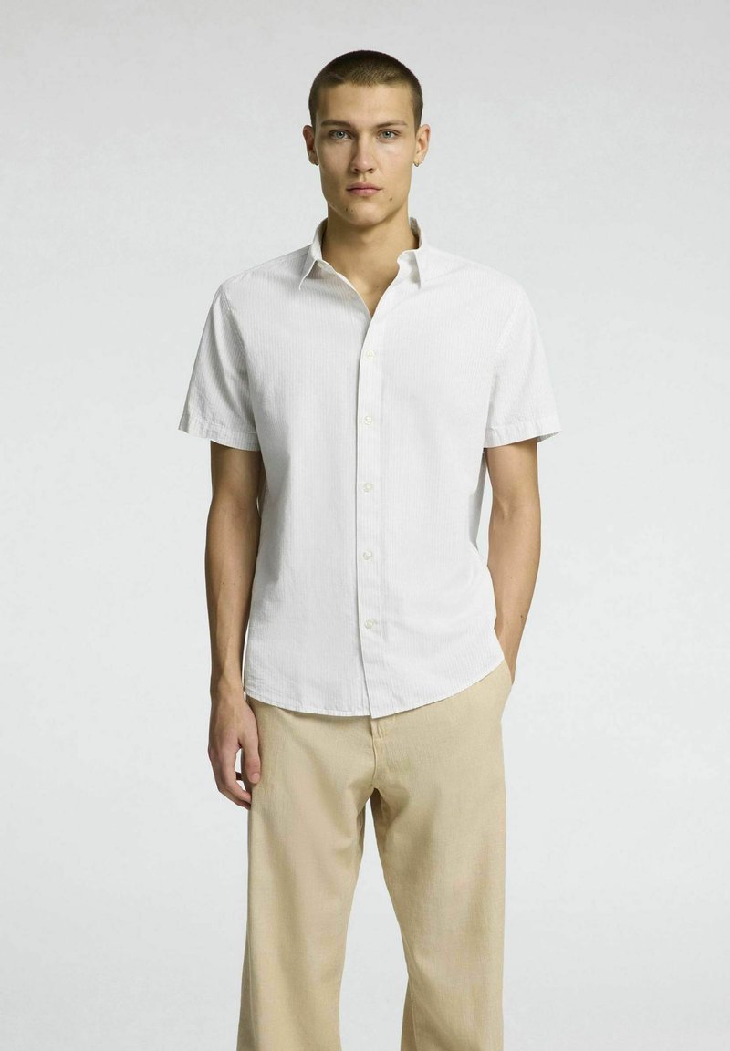 Kortärmad skjorta med knappar i vitt med ett subtilt randigt mönster, tillverkad av lätt tyg; kombinerad med ljusbeiga byxor.