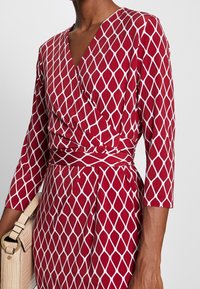 Robe portefeuille rouge à motif losange blanc, avec des manches trois-quarts et une taille cintrée. Accompagnée d'un sac beige avec des accents texturés.