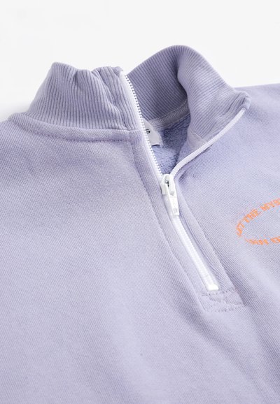 Sweat-shirt violet clair à fermeture éclair quart de zip avec col montant côtelé et petit texte orange partiellement visible sur la poitrine.