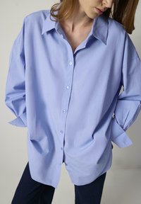 Cămașă lungă, cu mâneci lungi, de culoare albastru deschis, cu guler ascuțit și croială lejeră. Are un luciu subtil și prezintă mâneci suflecate și buzunare laterale.