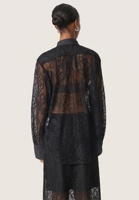 Camicia nera in pizzo con motivo floreale semi-trasparente, dotata di colletto classico e maniche lunghe con polsini a bottone.