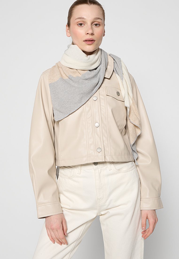 Kobieta w beżowej skórzanej kurtce na krótko, wielobarwnej szarej i beżowej szalu oraz off-white spodniach na tle jednolitym.