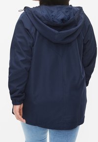 Kvinna som bär en mörkblå huva jacka med elastiska ärmslut, sedd bakifrån, i kombination med ljusblåjeans mot en neutral bakgrund.