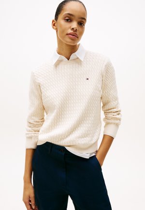 Vrouw draagt een crèmekleurige kabelgebreide trui over een witte blouse met kraag en donkerblauwe broek, staand met één hand in de zak.