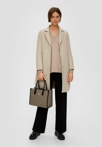 Beige jas over een lichtbruine trui, gecombineerd met zwarte wijde broek. Houdt een gestreepte beige en zwarte handtas met zwarte handvatten vast.