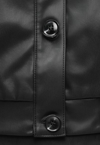 Crna kožna tkanina glatke teksture, s vertikalnom krpom i dva sjajna crna gumba s četiri rupe. Suptilni detalji šivanja su vidljivi.