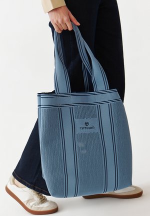 Blauwe netstof draagtasje met donkere strepen, voorzien van een logo, stevige handvatten en een gestructureerde afwerking. Draagt met donkere broek en lichte schoenen.