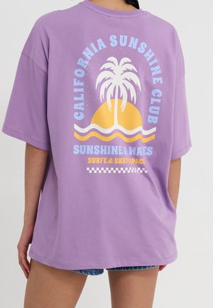Osoba nosząca fioletową oversize koszulkę z palmą, słońcem, falami i napisem "California Sunshine Club" oraz "Surfe & Skatespace" na plecach.