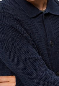 Marinblå stickad cardigan med en strukturerad mönster, knäppning med knappar och krage. Har långa ärmar med ribbad detalj på axlarna.