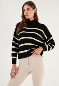 Zwart en crèmekleurig gestreepte gebreide pullover met een hoge hals, een relaxte pasvorm en ribbeltextuur, gecombineerd met lichtgekleurde leggings.