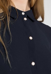 Navy blue blouse with a collar, featuring three decorative buttons with a pearl center and gold accents. Smooth, textured fabric.
