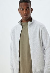 Grijze ritsjack met een hoge kraag, voorzien van een gladde textuur. Draagt over een olijfgroen shirt met korte mouwen. Schoon, minimalistisch ontwerp.