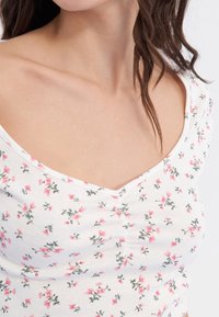 Mujer vestida con una blusa blanca con pequeños patrones florales en rosa y un escote fruncido, con mechones de pelo castaño sueltos sobre su hombro.