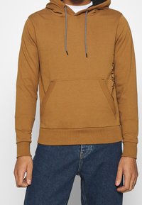 Sweat à capuche marron avec une poche kangourou, une capuche ajustable, et des cordons noirs. Présente une texture lisse ainsi que des poignets et un ourlet côtelés.